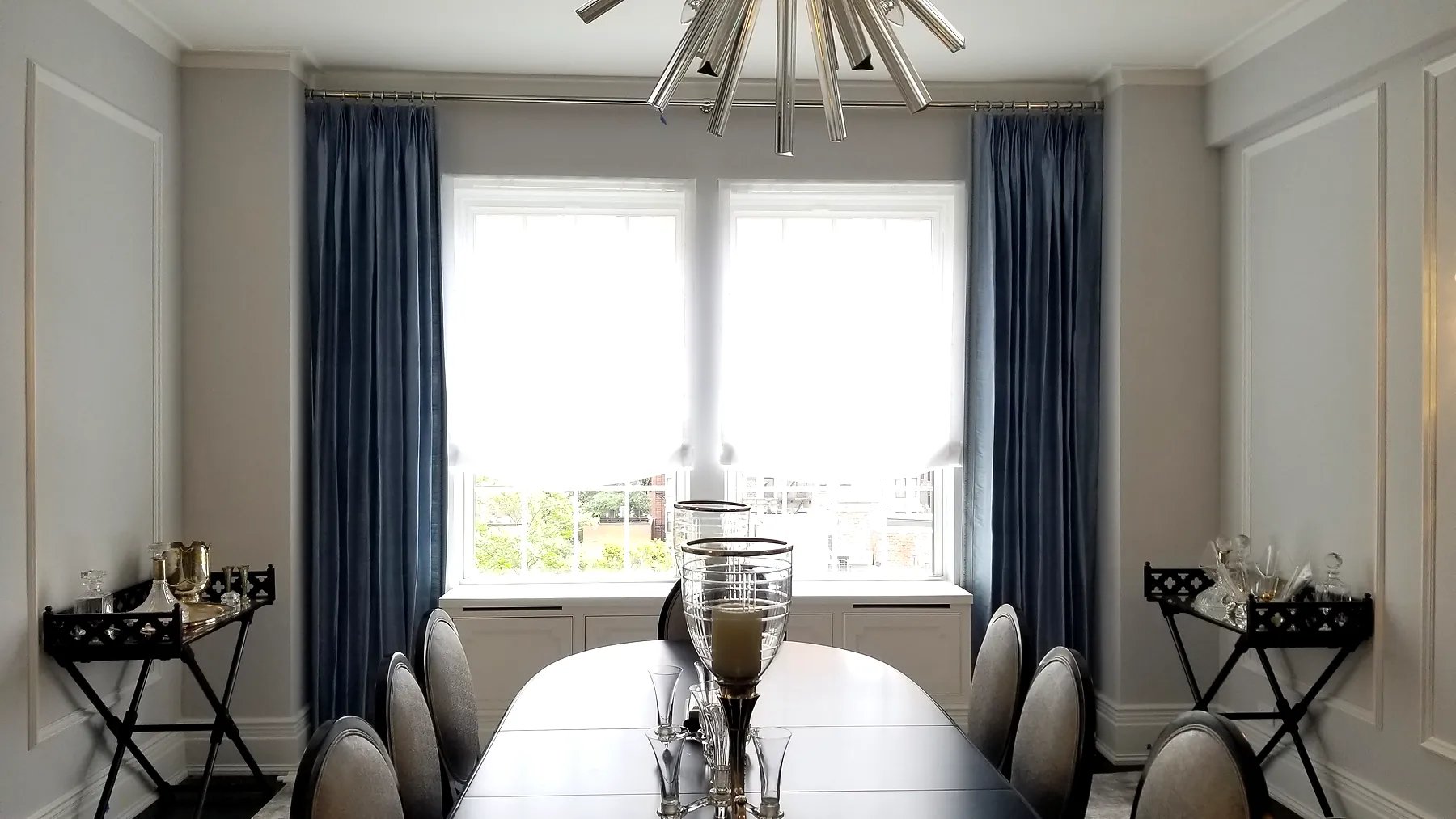 Pinch pleat drapery over paired dining room windows layered with roller shades